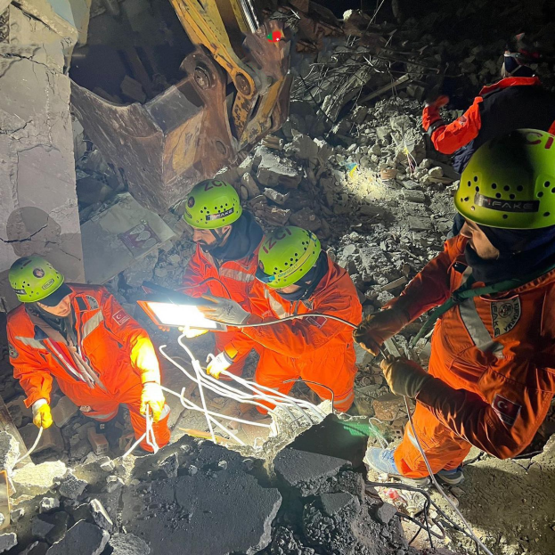 Gruppe von Menschen in orangefarbenen Uniformen und grünen Helmen bei der Arbeit an Schutt mit einem Kran im Hintergrund während eines Einsatzes nach dem Einsturz eines Gebäudes.