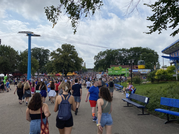 Eine Menschenmenge geht eine Straße mit Bänken, Grönflächen und urbanen Elementen entlang unter einem bewölkten Himmel, einige tragen Taschen in der Nähe eines Festivals.