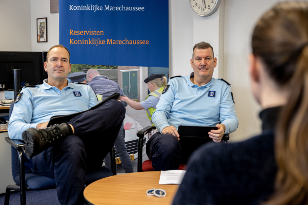 Zwei Polizeibeamte sitzen an einem Tisch mit einer Frau, Papieren und Gegenständen auf dem Tisch, einer Wandtafel und einer Uhr im Hintergrund, einem zweiten Tisch mit einem Monitor, einer Tastatur, einer Maus und Gegenständen links daneben und Fotorahmen an der Wand.