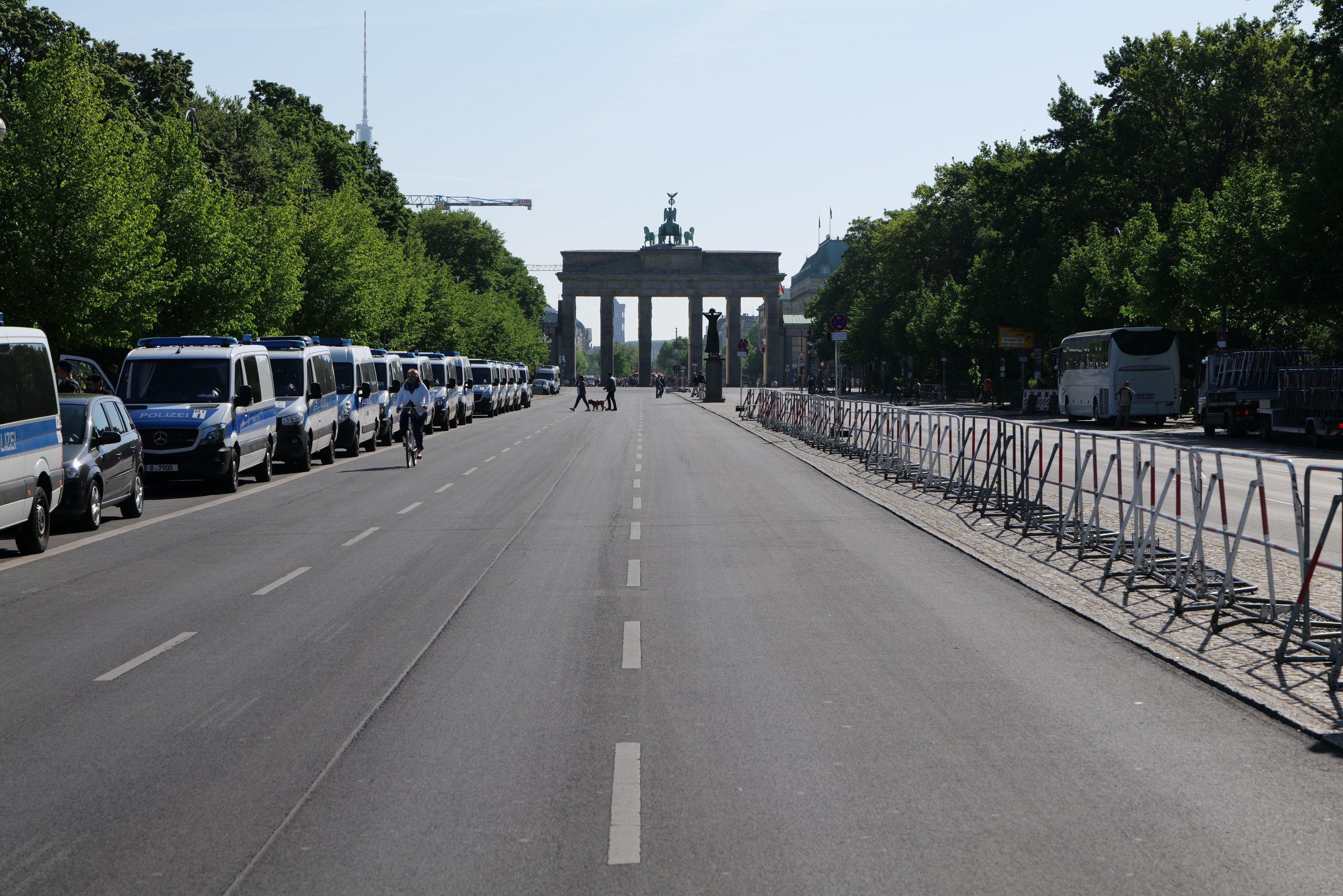 Lange Reihe von Polizeiwagen, die entlang einer Straße vor dem Brandenburger Tor geparkt sind, mit Fahrradfahrern, Füüßgängern, Absperrungen und Bäumen; ein Bogen mit Statuen im Hintergrund unter einem sichtbaren Himmel.