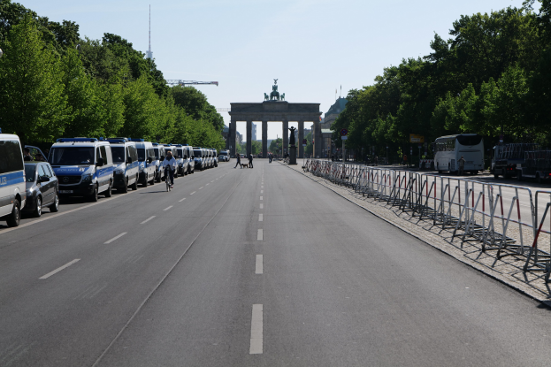 Lange Reihe von Polizeiwagen, die entlang einer Straße vor dem Brandenburger Tor geparkt sind, mit Fahrradfahrern, Füüßgängern, Absperrungen und Bäumen; ein Bogen mit Statuen im Hintergrund unter einem sichtbaren Himmel.
