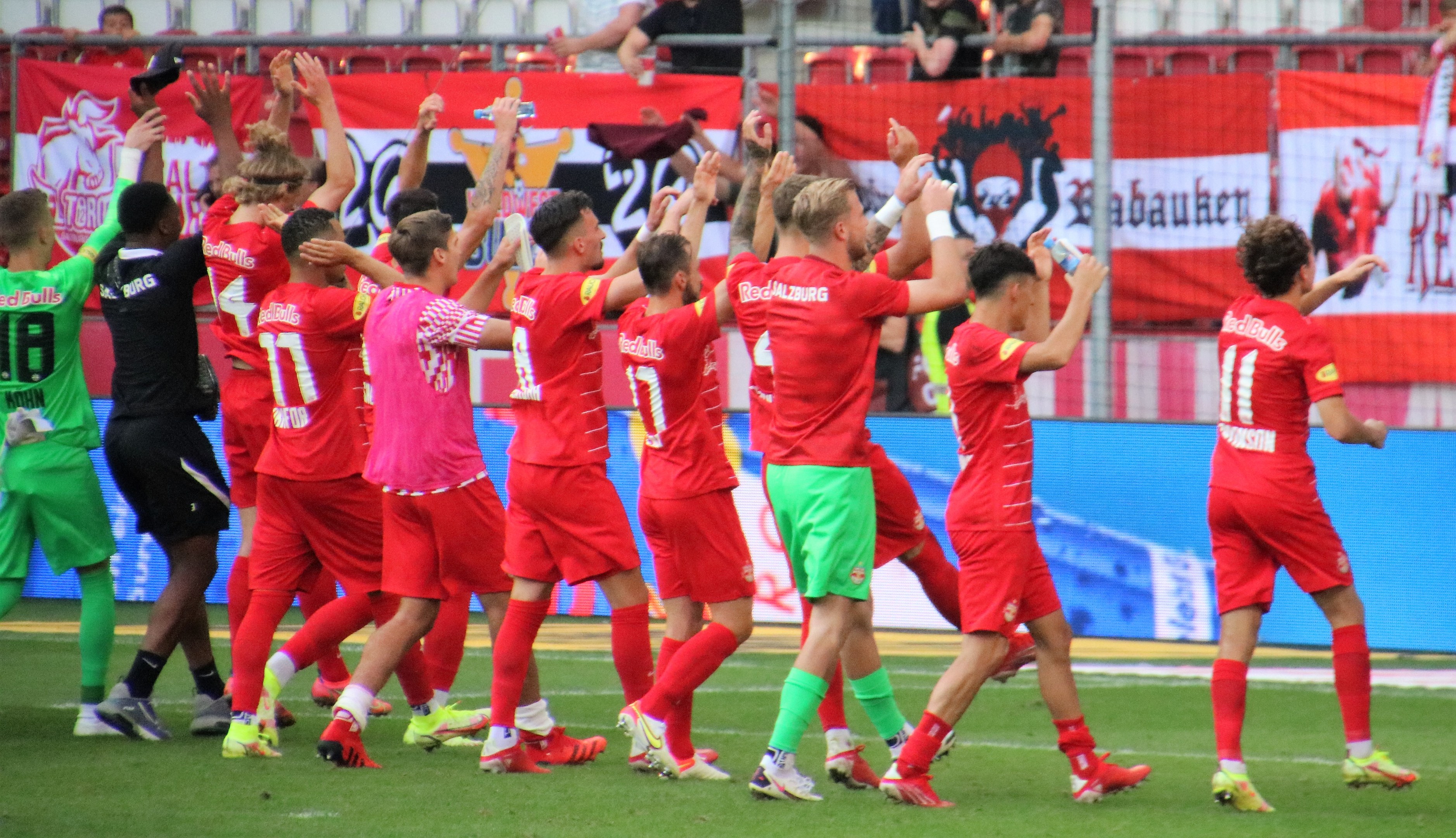 Gruppe von Fußballspielern, die ein Tor feiern, auf einem Feld, einige halten Flaschen oder klatschen, mit einem Zaun, Bannern, Metallstangen, Stühlen und einer Decke mit Deckenleuchten im Hintergrund.