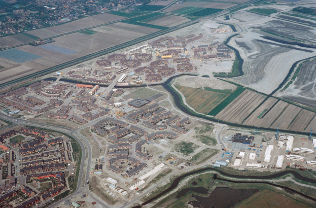 Luftaufnahme einer Stadt mit dichtem Baumbestand, Straßen, Feldern, Bäumen und einem zentralen Reservoir, umgeben von städtischen und grünen Bereichen.