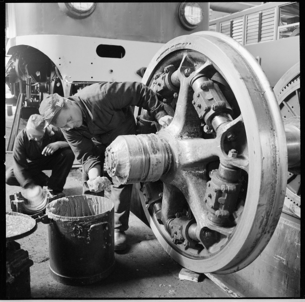 Zwei Männer mit Mützen arbeiten an einem Zugrad in einer Schwarz-Weiß-Fabrikumgebung, mit Werkzeugen und einem Eimer auf dem Boden und einem Fahrzeug im Hintergrund.
