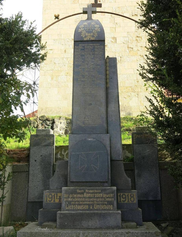 Denkmal vor einer Kirche mit Kreuz, gewidmet deutschen Soldaten, die im Zweiten Weltkrieg starben, umgeben von Bäumen und einer Mauer.