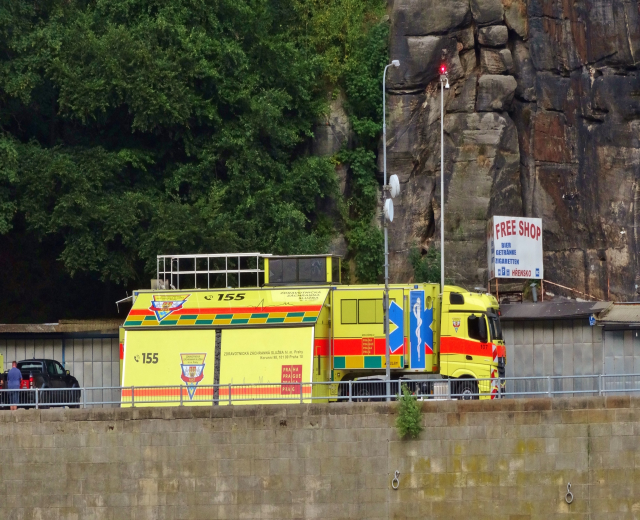 Ambulanz am Rande einer Straße neben einer Klippe mit verschiedenen Gegenständen im Vordergrund und Bäumen und einem Auto im Hintergrund.