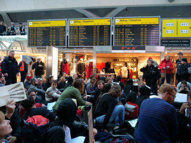 Eine große Gruppe von Menschen sitzt in einem Flughafen, einige mit Taschen und Papieren, andere stehen, mit Schildern, Puppen in Kleidern und Deckenlichter im Hintergrund, was auf eine Demonstration hindeutet.