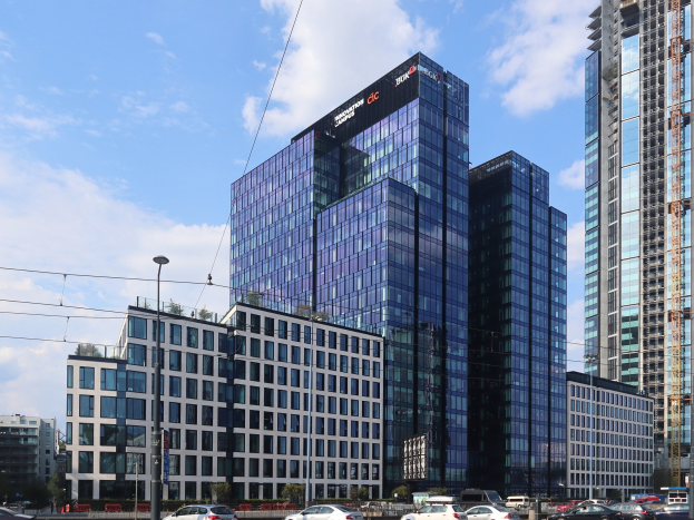 Großes Hauptquartier-Gebäude der Deutschen Bank in Frankfurt, Deutschland, mit vielen Fenstern, umgeben von städtischer Infrastruktur einschließlich Straßenlaternen, Fahrzeugen, Grünflächen und einem bewölkten Himmel.