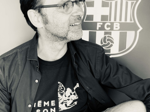 Ein Mann mit Brille und schwarzem Hemd sitzt vor einem schwarz-weißen FC Barcelona-Logo auf einer Wand und lächelt.