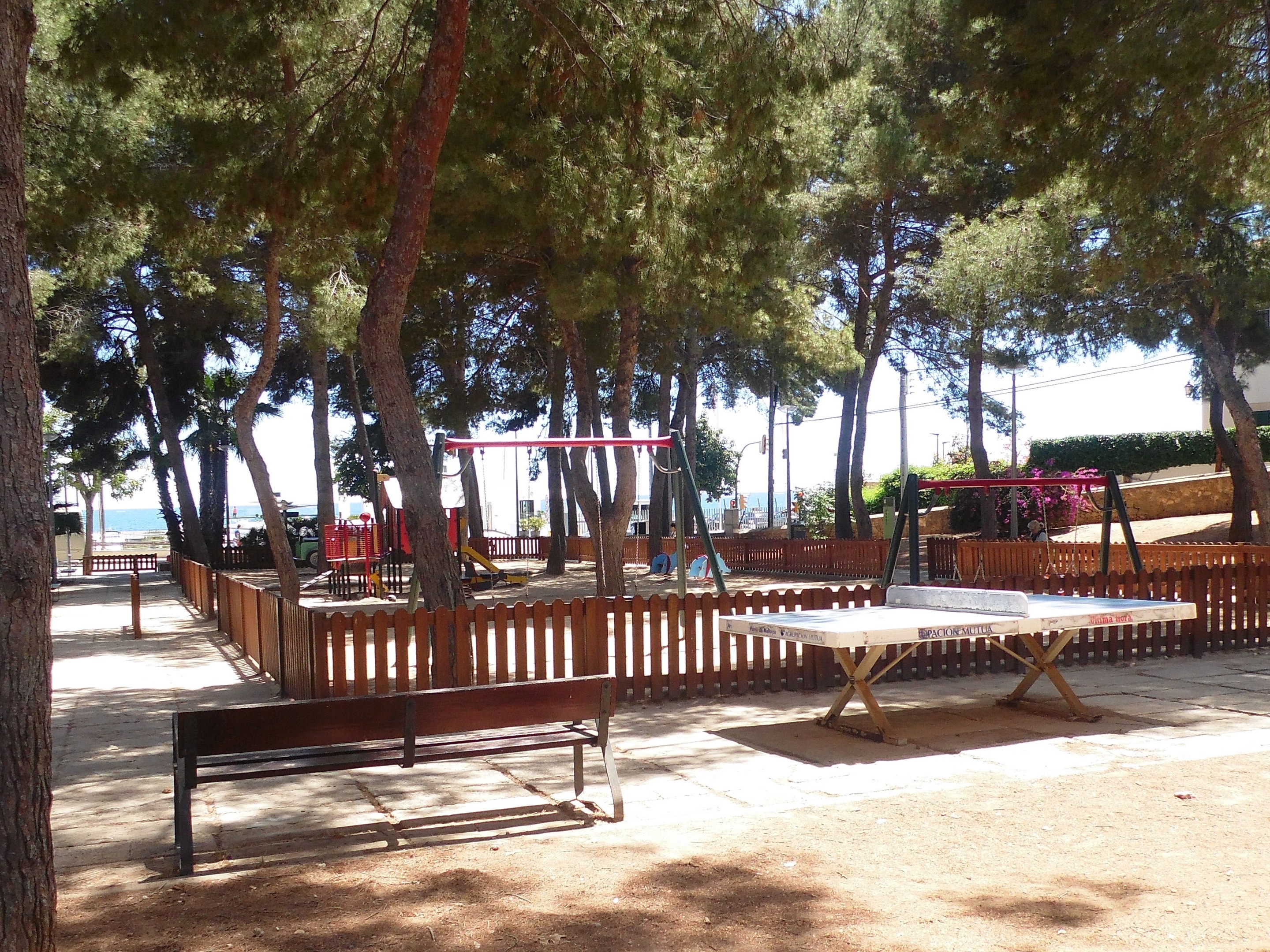 Ein Park mit einem zentralen Tischtennis-Tisch, umgeben von Bänken, Schaukeln, Bäumen und anderen Gegenständen, mit Blick auf einen See unter einem klaren blauen Himmel.