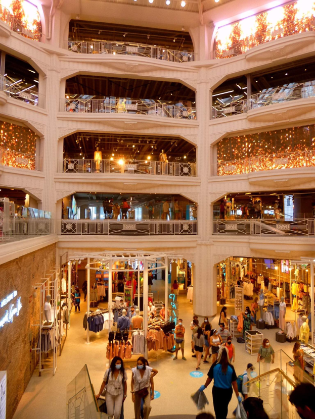 Ein belebter Einkaufszentrumsvorplatz mit Menschen, die Taschen tragen, Schaufensterpuppen in Kleidern, Geländern, Deckenleuchten und Texttafeln an den Wänden.