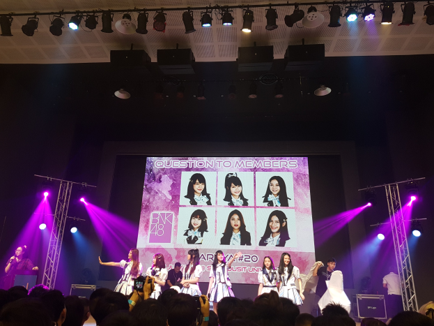 Eine Gruppe von Menschen, die auf einer hell erleuchteten Bühne performen, mit einer Menge darunter, einem Menschen mit Mikrofon und einem anderen, der ein Instrument spielt, und einem Bildschirm mit Bildern der K-Pop-Gruppe BK48 im Hintergrund.