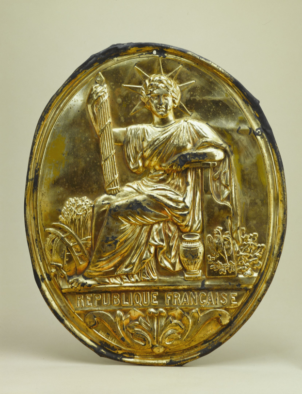 Goldene runde Plakette mit einem reliefierten Abbild der Freiheitsstatue und eingraviertem Text auf weißem Hintergrund.