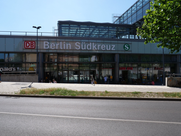 Ein großes Glaswand-Bahnhof namens "Berlin Südkreuz" in Berlin, Deutschland, mit Straßenlaternen, Lichtern, Fahrzeugen, Fußgängern, Fahrrädern, Bäumen und einem klaren blauen Himmel.