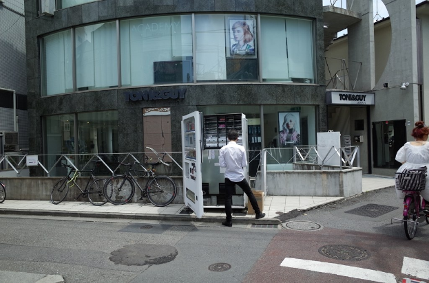 Eine Straße mit einer Frau auf einem Fahrrad, einem Mann in der Nähe einer Bude und mehreren Fahrrädern in der Nähe eines Ladens.