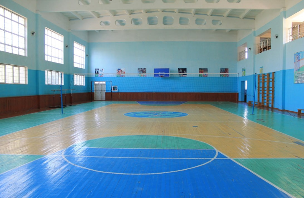 Große Turnhalle mit blau-weißem Boden, Basketballfeldmarkierungen, einem Netz mit Pfosten, einer Tür, Fenstern, Wandplakaten, einem Geländer und einem Dach.