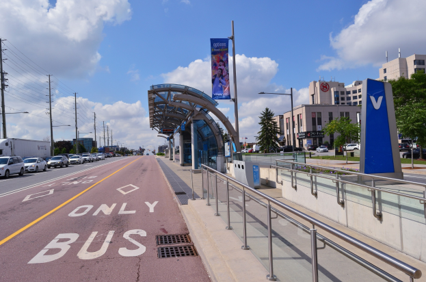 Stadtstraße mit parkenden Autos, eine Bushaltestelle mit Geländern und einem Textschild auf der rechten Seite, flankiert von Gebäuden, Bäumen, Laternen und Oberleitungen.