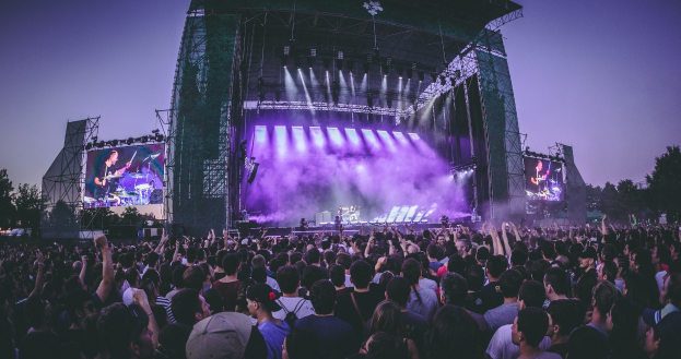 Eine große Menge bei einem Musikfestival schaut auf einer hell erleuchteten Bühne mit Instrumenten, Bildschirmen und einer Kulisse aus Bäumen, einem Gebäude und einem klaren blauen Himmel.
