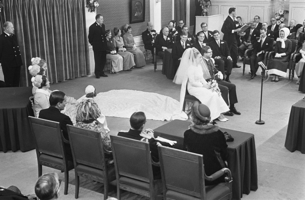Schwarzes und weißes Foto einer Hochzeitszeremonie mit sitzenden und stehenden Menschen, einem Mann mit Mikrofon, einer Frau mit einem Blumenstrauß, einem Podium mit Mikrofon auf der rechten Seite und Blumengestecken und einem Vorhang auf der linken Seite.