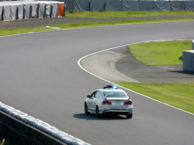 Polizeiauto fährt auf einer Rennstrecke mit Gras auf beiden Seiten, Menschen und Zelte sind im Hintergrund sichtbar.