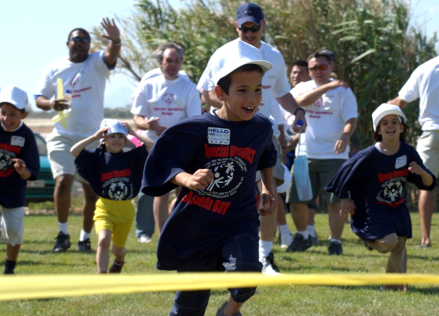 Kinder bei einem Lauf auf einer grünen Wiese mit einem gelben Band im Vordergrund, einige tragen Kappen und Schutzbrillen, andere halten Gegenstände, vor einem klaren blauen Himmel mit Bäumen im Hintergrund.