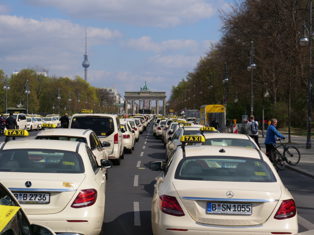 Eine lange Reihe von Taxis steht entlang einer Straße in Berlin, Deutschland, mit fahrenden Fahrzeugen, Radfahrern, Fußgängern, Laternenpfählen, Bäumen, Gebäuden, einem Bogen und einem Turm im Hintergrund unter einem bewölkten Himmel.
