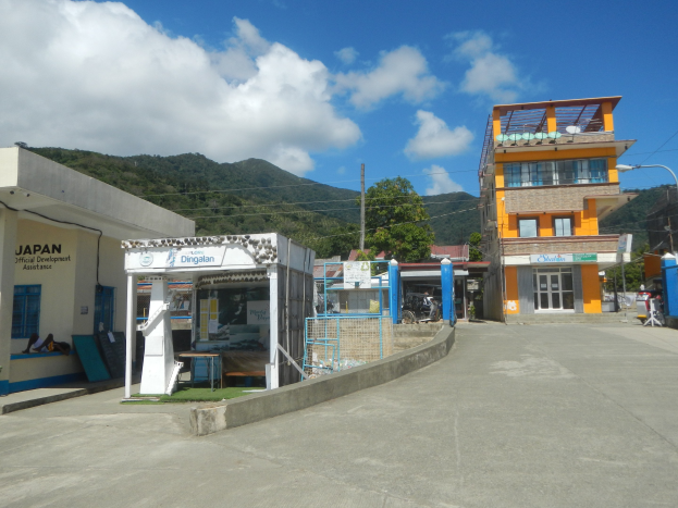 Kleinstadt-Szene mit einer Tankstelle im Vordergrund, parkende Fahrzeuge, umgeben von Gebäuden, Straßeninfrastruktur, Bäumen, Hügeln und einem bewölkten Himmel.