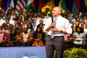 Präsident Obama spricht auf einer Wahlveranstaltung, hält ein Mikrofon und steht vor einer Menge von Menschen, einige sitzen und einige stehen, sowie einer Pflanze im Vordergrund, Fahnen und eine Tafel mit Text im Hintergrund.