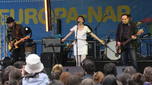 Vier Musiker treten auf der Bühne auf: Eine Frau in einem weißen Kleid singt in der Mitte, flankiert von zwei Männern, die Gitarren spielen, während eine Menge darunter eine Wand mit Lautsprechern im Hintergrund zusieht.