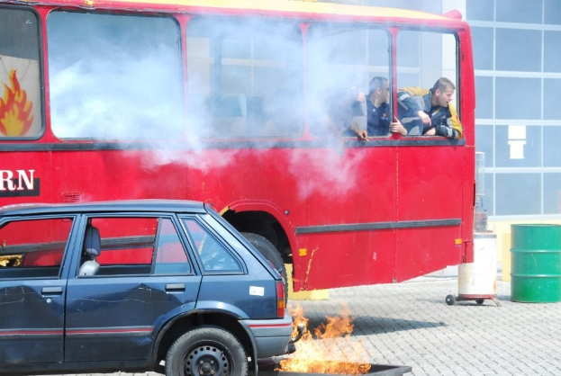 Roter Doppeldeckerbus mit Rauch, daneben geparktes Auto, drei sichtbare Passagiere im Bus und ein Gebäude mit Glasfenstern und einem Fass im Hintergrund.