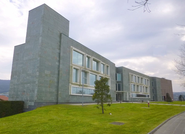 Großes Gebäude mit grünem Rasen, Bäumen, Pflanzen und einer Straße davor, umgeben von Hügeln und einem bewölkten Himmel, beschriftet als Universität Stuttgart.
