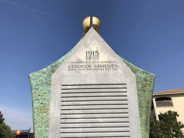 Ein steinernes Denkmal mit einer Uhr darauf, umgeben von Bäumen und Gebäuden, mit einer Inschrift, die den 24. Jahrestag des Völkermords in Armenien gedenkt.
