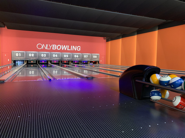 Eine Bowlingbahn mit Bowlingkugeln rechts, Pins in der Mitte, einer textbedeckten Wand im Hintergrund und einer sichtbaren Decke.