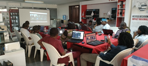 Gruppe von Menschen um einen Tisch mit Laptops und anderen Gegenständen während eines digitalen Marketing-Workshops, mit einem Banner, einem Display und Deckenleuchten im Hintergrund.