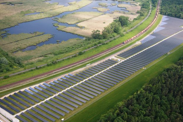 Luftaufnahme eines Solarparks mit Panelen, umgeben von Bäumen, Gras, Wasser und einer nahen Bahnstrecke.