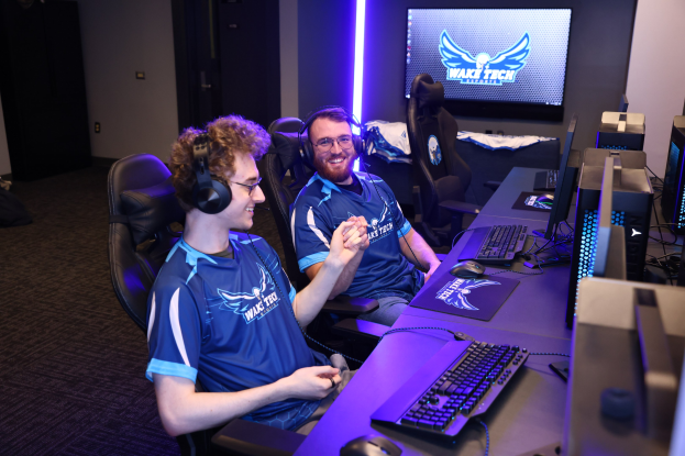 Zwei Männer in blauen T-Shirts und Kopfhörern sitzen an einem Schreibtisch mit Computerausrüstung und spielen ein Spiel in einem Raum mit einem Bildschirm und Lichtern, während Taschen auf dem Boden sichtbar sind.