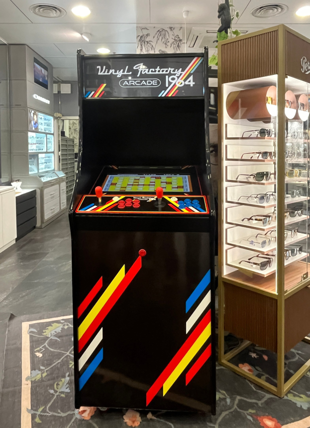 Ein Geschäft mit einem vintage Arcade-Automaten in der Mitte, einem Glasvitrine auf der rechten Seite und einer retro-stylischen Beleuchtung an der Decke mit Schränken und einem Fernseher im Hintergrund.
