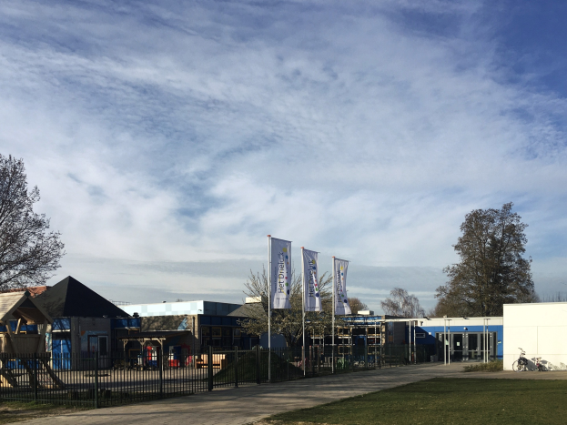 Großes Gebäude mit Metallzaun, Weg, Bäumen, Bannern, einem geparkten Fahrrad, nahen Häusern und einem bewölkten Himmel, identifiziert als der neue Spielplatz für Kinder an der Schule.