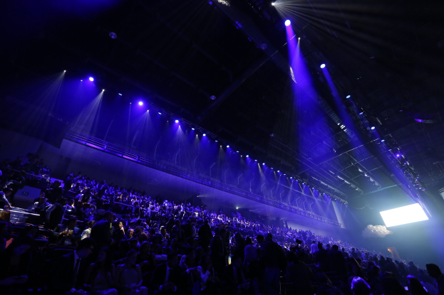 Eine große Menschenmenge, die in einem Konzertsaal auf einer Bühne sitzt, mit Deckenbeleuchtung und einem Bildschirm auf der rechten Seite.