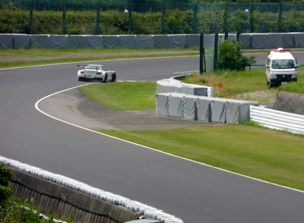 Rennwagen auf einer Rennstrecke mit einem Polizeiwagen im Hintergrund, umgeben von Gras, Pflanzen, Bäumen und Zäunung, mit einem Fahrzeug auf der rechten Seite und Text am unteren Rand.