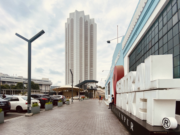 Ein belebter zentraler Markt in Kuala Lumpur, Malaysia, mit hohen Gebäuden, Straßenlaternen, Fahrzeugen, Fußgängern, Schildern, Bäumen und einem bewölkten Himmel.