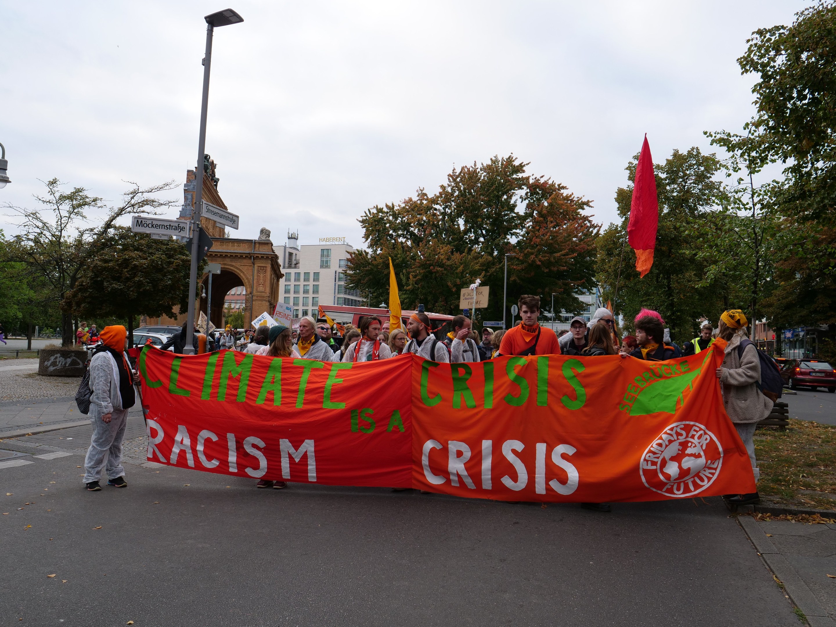 Eine Gruppe von Menschen marschiert eine von Bäumen gesäumte Straße entlang und hält ein Banner hoch, auf dem "Klima-Krise ist eine Krise" steht, mit Fahrzeugen und Gebäuden im Hintergrund unter einem klaren blauen Himmel.