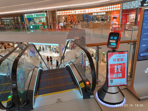 Eine Rolltreppe in einem Einkaufszentrum mit Menschen, die nach unten gehen, ein Bildschirm mit Text auf der rechten Seite und verschiedene Geschäfte und Gegenstände im Hintergrund.