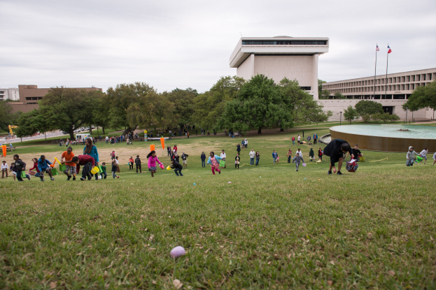 Gruppe von Menschen, die mit Ostereiern in einem grünen Feld mit Bäumen, Gebäuden, Fahnen, einem Brunnen und einem bewölkten Himmel spielt.