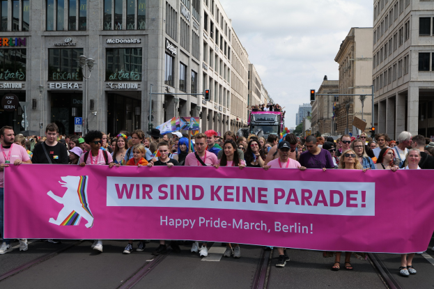 Gruppe von Menschen, die eine Straße in Berlin entlanggehen und eine pinkfarbene "Happy Pride March"-Fahne halten, mit Gebäuden, Laternenmasten und Verkehrszeichen an der Straße.