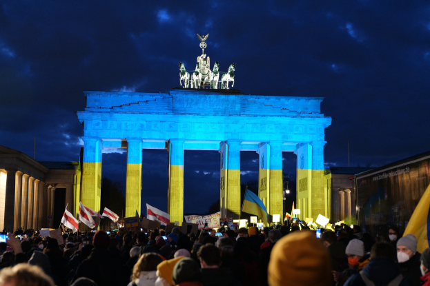 Menschenmenge mit Fahnen und Schildern vor dem Brandenburger Tor, mit einem Banner auf der rechten Seite mit protestbezogenem Text.