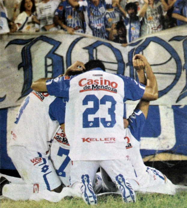 Eine Gruppe von Fußballspielern feiert ein Tor auf dem Boden vor einer jubelnden Menge, mit einem Banner im Hintergrund.