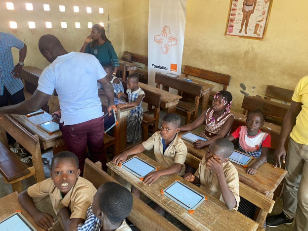 Gruppe von Kindern an Tischen sitzend, die Tablets verwenden, in einem Klassenzimmer mit Erwachsenen in der Nähe, einem Banner und einem Plakat an der Wand.