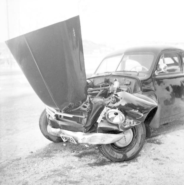 Ein stark beschädigtes Schwarz-Weiß-Foto eines an einem Crash beteiligten Autos mit eingedellter Haube, zersplitterten Fenstern und einer offenen Fahrertür, die den Motor freigibt.