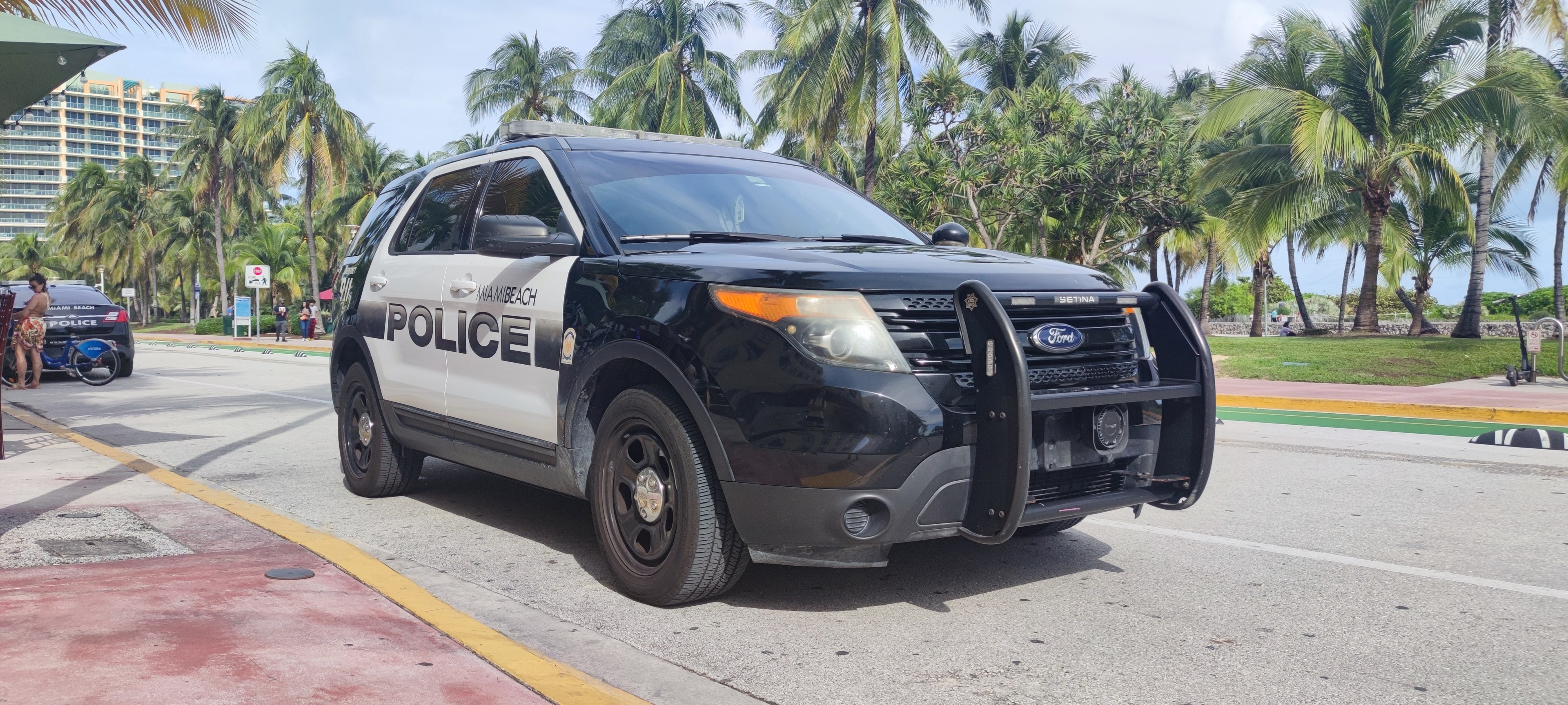 Polizeiauto auf dem Seitenstreifen geparkt mit Menschen, Fahrrädern, Gras, Bäumen, Gebäuden und einem klaren blauen Himmel im Hintergrund; das Vorderrad ist sichtbar.